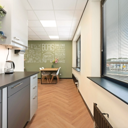 Kantoorruimte huren op Stationsplein 4, Zwijndrecht. Moderne pantry met lichtinval via grote ramen, uitgerust met keukenkastjes, beklede vloer en stoelen. Ideaal voor zakelijke pauzes en informele vergaderingen.