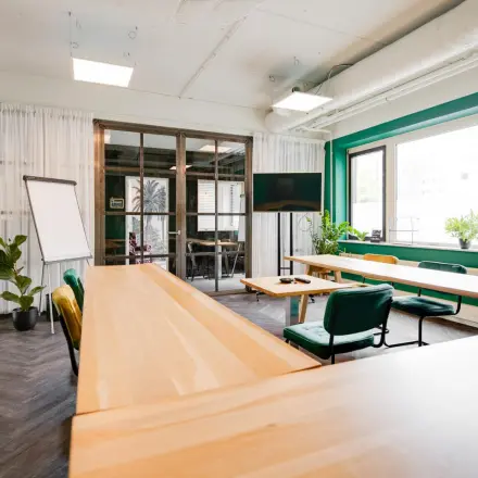 Kantoorruimte huren op Hanzeplein 11-27 in Zwolle met houten tafels, een flip-over en een groot scherm. Groen-witte tinten met veel lichtinval, ideaal voor vergaderingen. Planten voegen een frisse sfeer toe.