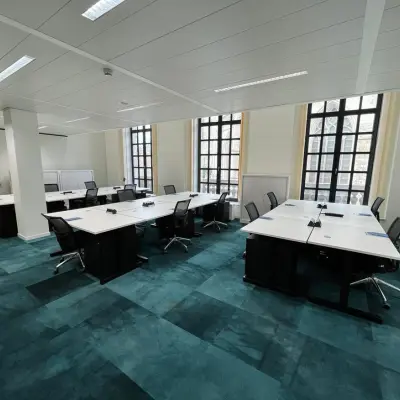 Ruime kantoorruimte te huur op Rue des Colonies 56, Brussel Europese Wijk, met grote ramen en blauwe vloertapijten. Verschillende bureaus en stoelen klaar voor gebruik in een lichte omgeving.