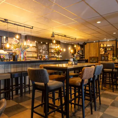 Cozy lounge area with high tables and barstools, surrounded by warm lighting and decorative elements, ideal for informal meetings. Consider this inviting space for office space rental in Almelo.