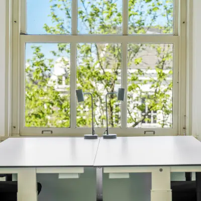 Bright office space with two desks and chairs, complemented by a large window overlooking greenery, located in Amsterdam Canal Belt, perfect for office space rental.