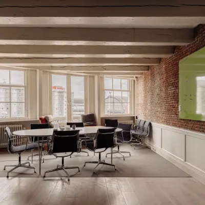 Charming office space at Prins Hendrikkade 14 in Amsterdam Center with exposed brick walls, a large table, modern chairs, and a large window with city views. Ideal for office space rental.