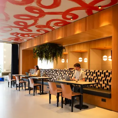 Bright office space with modern seating areas, artistic ceiling design, and individuals engaged in work at Barbara Strozzilaan 101, Amsterdam Zuidas. Ideal for rent office space.