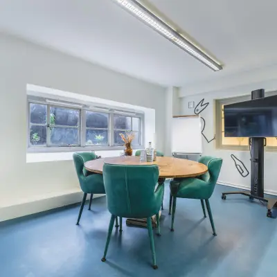 Bright office space in Breda Centrum-Station, featuring a round wooden table with green chairs, a flat-screen TV, and a whiteboard, ideal for office space rental.