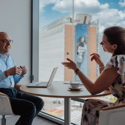 Two business professionals engage in a discussion at a round table in a modern office with a view of Brussels European District, ideal for office space rental.