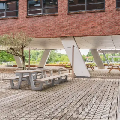 Outdoor view of Rijnzathe 12 in De Meern, featuring wooden picnic tables under a shaded area with modern architecture.