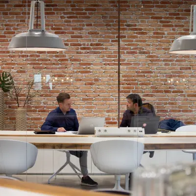 Two people engaged in discussion at a modern, furnished workspace with exposed brick walls in Eindhoven Strijp-S. Ideal for office space rental seekers.