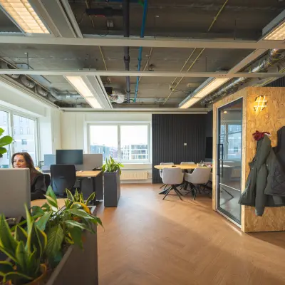 Modern office space at Brouwerijstraat 1, Enschede with contemporary furnishings, plants, and a glass-walled meeting area. A person is working at a desk, showcasing available office space rental options.