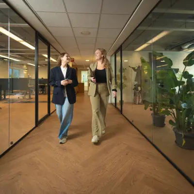 Two people walking and conversing in a modern office to rent at Brouwerijstraat 1, featuring glass walls and wooden flooring with office plants.