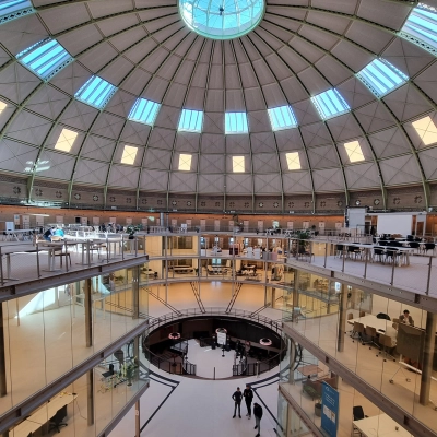 Spacious office to rent at Koepelplein 1E, Haarlem Centrum. Featuring a dome ceiling with abundant natural light, furnished workspaces and people engaged in discussions, ideal for office space rental.