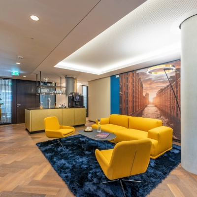 Welcoming lounge area at Alter Wall 32, Hamburg Old Town, featuring modern yellow seating, a blue rug, and a large cityscape mural, ideal for office space rental.