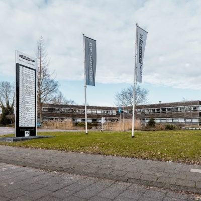 Exterior view of an office building at De Vos van Steenwijklaan 75, Hoogeveen, surrounded by greenery with signage and flags, offering office space rental options.