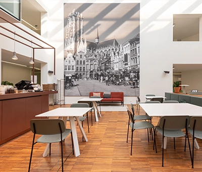 Bright office space in Mechelen Centrum with modern furniture and a large mural, ideal for office space rental. Tables and chairs arranged for collaborative work.