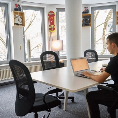 Man working on a laptop in a Munich Giesing office space, featuring a round table, several chairs, and decorated with skateboards by the windows.