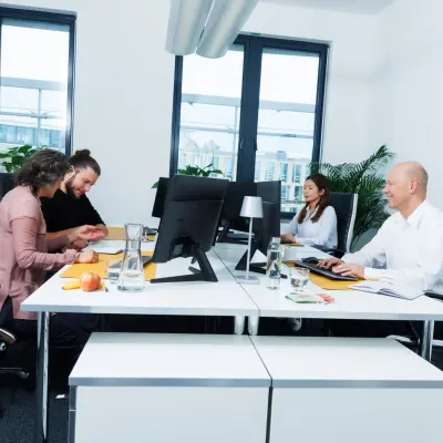 People collaborating in a modern, furnished Munich Laim office space with computers and documents on white desks, available for office space rental.