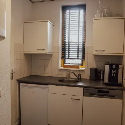 Compact kitchenette with white cabinets, small fridge, coffee machine, and a window with blinds in an office space to rent.