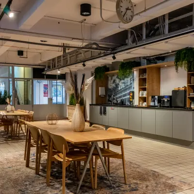 A modern office space at Van Nelleweg 1, Rotterdam Delfshaven, featuring wooden tables, chairs, potted plants, and a kitchenette, ideal for office space rental.