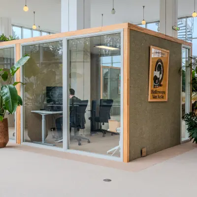 Office space rental at Van Nelleweg 1, Rotterdam Delfshaven. A person works at a desk inside a glass-walled office unit, surrounded by plants and modern furnishings.