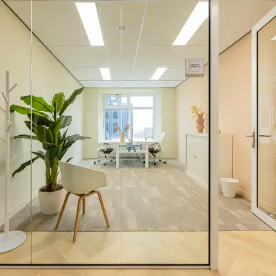 Bright office space with glass walls, two desks, chairs, and green plants in Rotterdam's Kop van Zuid, ideal for rent office space.