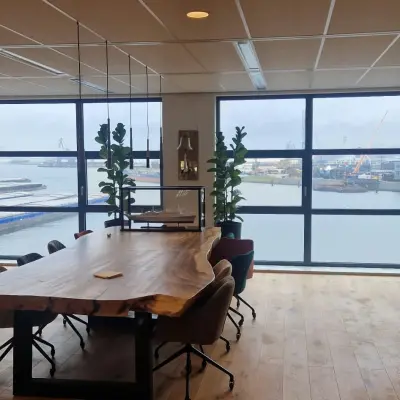 Modern office space with a large wooden table and chairs, overlooking Rotterdam Waalhaven through expansive windows.