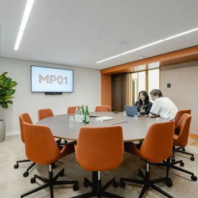 A modern conference room at Moreelsepark 1, Utrecht Central Station with a round table, orange chairs, a large plant, and two people discussing a project. Ideal for office space rental.
