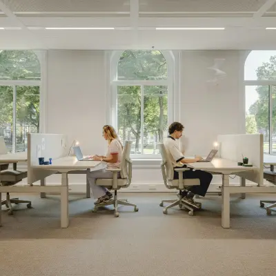 People working at desks in a bright, well-lit office space with large windows in Utrecht Central Station, ideal for office space rental.