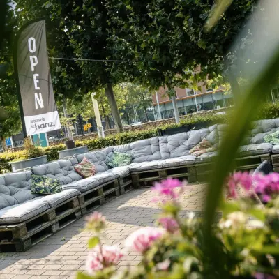 Outdoor seating area with cushioned benches made from pallets, surrounded by greenery, at Hanzeplein 11-27, Zwolle. Ideal for a break or informal meeting. Office space rental nearby.