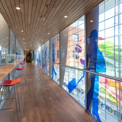 Moderne gang met glazen wanden en kleurrijke decoraties in kantoorpand op Stationsplein 49, Amersfoort Centraal Station. Hoge barkrukken langs houten tafel bieden uitzicht op buiten.