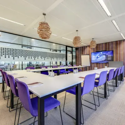 Vergaderruimte met paarse stoelen en grote tafels in een modern kantoorgebouw aan Prins Bernhardplein 200, Amsterdam Amstel. Ruime opzet met scherm en stijlvolle verlichting.