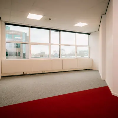 Ongemeubileerde kantoorruimte op Overschiestraat 182, Amsterdam, met lichtinval door grote ramen en een grijs met rood tapijt. Ideaal voor jouw nieuwe kantoorpand huren in De Schinkel.
