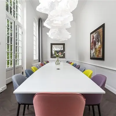 Vergaderruimte in kantoorpand op Keizersgracht 62-64 met lange tafel en kleurrijke stoelen, omringd door grote ramen en moderne kunst aan de muur. Kantoorruimte huren in Amsterdam Grachtengordel.