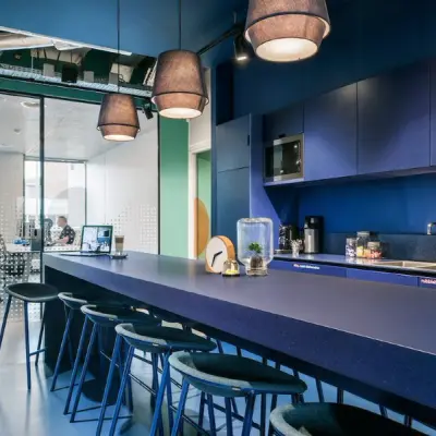 Moderne kantoorruimte aan de Jan van Galenstraat 335 in Amsterdam Nieuw-West, met een grote blauwe tafel en barkrukken, zicht op vergaderruimte. Twee mensen werken achter hun laptops.