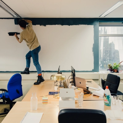 Een kantoor in Amsterdam Noord, TT Vasumweg 44, met een persoon die een muur schildert en bureaus die laptops, kantoorspullen en planten bevatten.