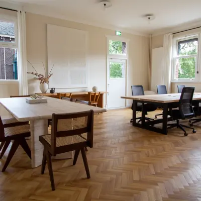 Ruime, lichte kantoorruimte met houten vloer en grote ramen aan de Emmalaan 7-9 in Amsterdam Oud Zuid. Verschillende bureaus en stoelen aanwezig voor huur.