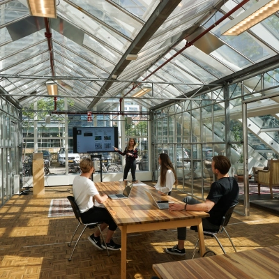 Kantoor met glazen wanden in Amsterdam Riekerpolder, gelegen op Johan Huizingalaan 400. Drie mensen bij vergadertafel, één presenteert aan een scherm. Ruimte biedt een heldere en moderne werkomgeving.