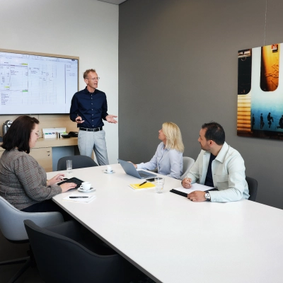 Zakelijke bijeenkomst in een moderne vergaderruimte op Teleportboulevard 110, Amsterdam Sloterdijk. Vier personen, waaronder een presentator, bespreken werkdocumenten bij een lange tafel met laptops en scherm. Kantoor huren mogelijk.