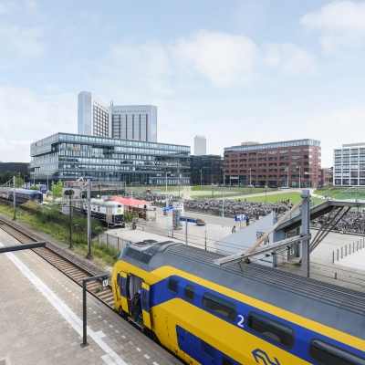 Buitenzijde van kantoorpand op Teleportboulevard 110 in Amsterdam Sloterdijk, met nabijgelegen treinsporen en een gele NS-trein. Ideale locatie voor kantoorruimte huren met uitstekende openbaar vervoer verbindingen.