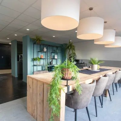 Een moderne kantoorruimte met lange houten tafel en gestoffeerde stoelen, groene planten en grote hanglampen. Ideaal om kantoorruimte te huren in Apeldoorn op Prins Willem-Alexanderlaan 401.
