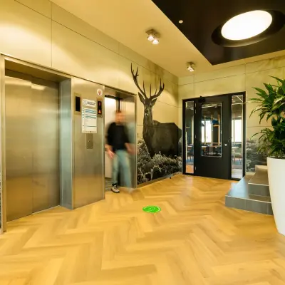 Hal met uitzicht op twee liften, houten vloer in visgraatpatroon, grote plantenbak en wanddecoratie met hertenafbeelding. Persoon loopt richting de deur. Geschikt voor kantoorruimte huren.