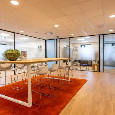 Moderne kantoorpand met glazen wanden en hoge houten tafels op een oranje tapijt. Ideale werkplek voor kantoorruimte huren in Bodegraven, Marktstraat 1-7.