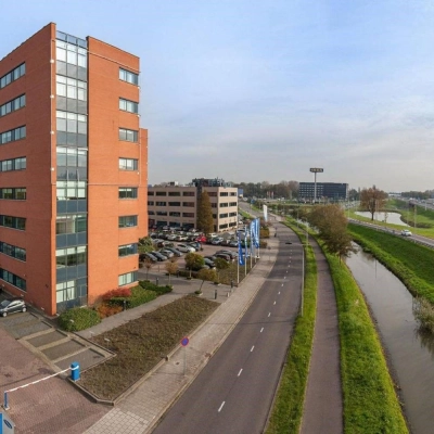 Kantoorpand aan de Olof Palmestraat 10 in Delft, met meerdere parkeerplaatsen voor het gebouw langs een rustige weg en een aangrenzende sloot.
