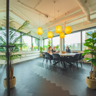 Lichte kantoorruimte met grote ramen, zes hanglampen en tafels met stoelen, gelegen op Hambakenwetering 1, Den Bosch. Twee personen zijn bezig met een zakelijke bespreking.