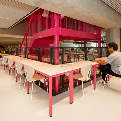 Moderne kantoorruimte in Den Haag Centrum met lange tafel, roze accenten en mensen die werken op laptops onder opvallende trappen. Geschikt voor kantoorruimte huren.
