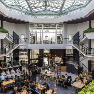 Ruime, gemeubileerde kantoorruimte met meerdere verdiepingen bij Eindhoven Airport, geschikt voor werk en overleg, met een glazen plafond voor veel natuurlijk licht. Mensen voeren zakelijke gesprekken.