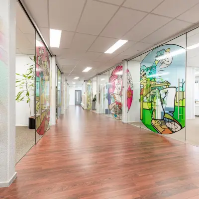 Kleurrijk kantoorpand op Meerenakkerplein 11-20 in Eindhoven De Hurk, met glaspanelen met kunstwerken langs de gang. Ideaal voor wie kantoorruimte wil huren met een inspirerende uitstraling.