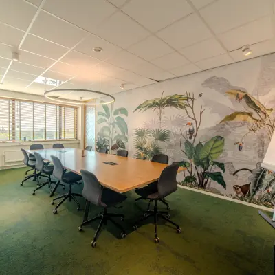Vergaderruimte met grote houten tafel en acht stoelen op Torenallee 20 in Eindhoven Strijp-S. De muren zijn versierd met een plantenmotief, ideaal voor wie een kantoorpand wil huren.