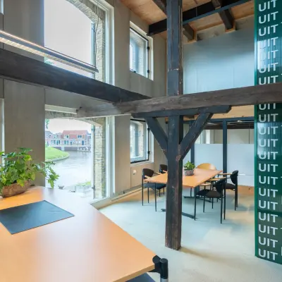 Kantoorruimte op de eerste verdieping van een kantoorpand aan Prins Hendrikkade 3 in Franeker, voorzien van houten balken, grote ramen, en diverse werkplekken met bureaus en stoelen.