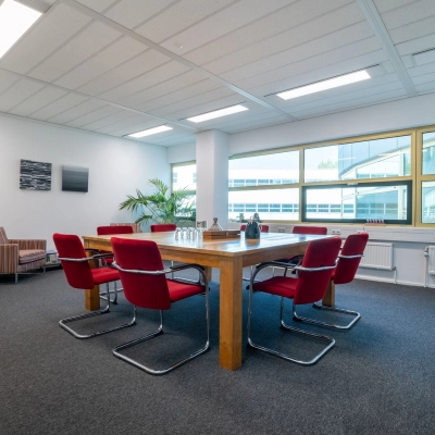 Ruime vergaderkamer in kantoorpand aan Groningenweg 8 8, Gouda, met rode stoelen rondom een houten tafel, grote ramen en kamersplanten. Ideaal voor kantoorruimte huren.