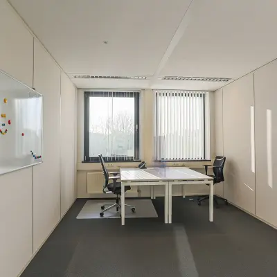 Lichte kantoorruimte met een wit bureau en twee zwarte stoelen, witte muren en een groot raam met verticale lamellen op Zernikepark 12, Groningen.