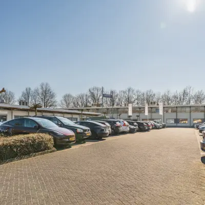 Parkeerplaats voor een kantoorpand aan de Steenovenweg 5 in Helmond, omringd door geparkeerde auto's en groene heggen onder een heldere blauwe lucht.